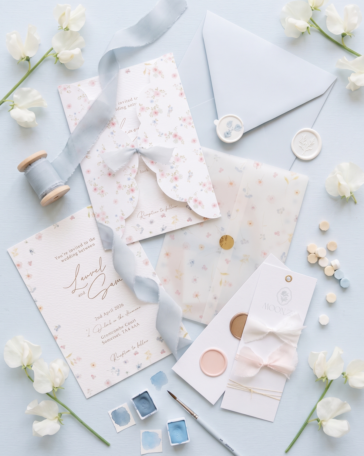 A styled flat lay of pastel stationery items on a light blue background, including a printed invitation card, envelope, ribbon, wax seals, paint swatches, a paintbrush, and small decorative flowers.