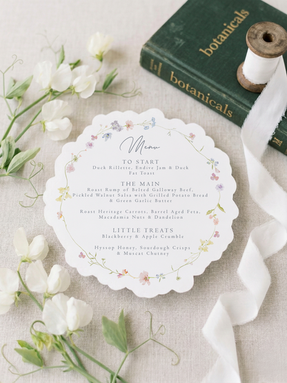 Menu card with floral design on a table with white flowers and a book titled 'botanicals'.